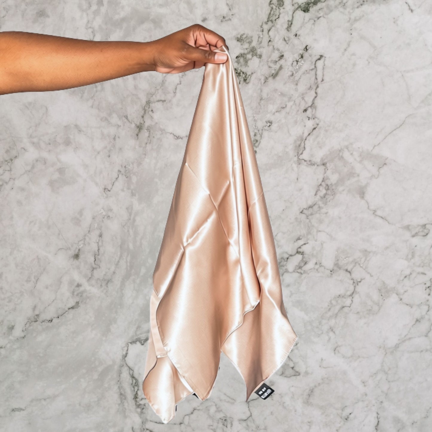 A person's hand holds up a square silk headscarf in a light champagne beige colour against a grey and white marble background.