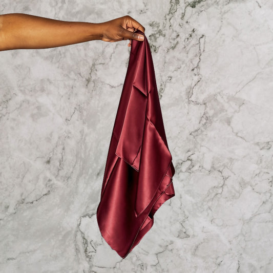 A hand holding up a chocolate brown silk hair scarf against a light grey marble background.
