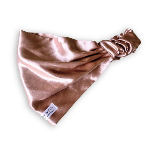 A satin headband in a dusty beige gold color, featuring a knotted back design and a small white brand tag that reads 'SLEEP WELL'.