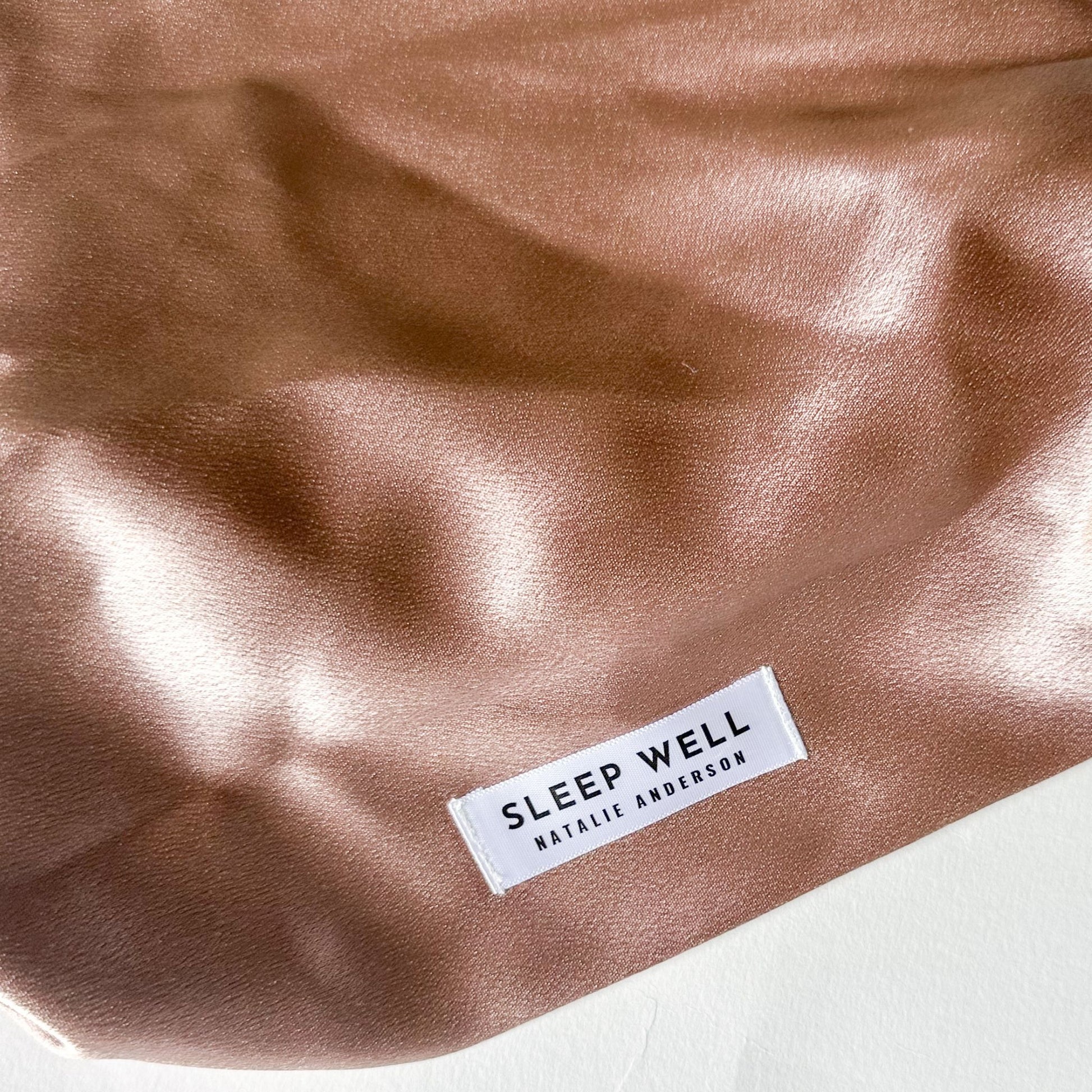 Close-up of a satin metallic beige gold fabric with a white woven label reading 'SLEEP WELL' and 'NATALIE ANDERSON'.